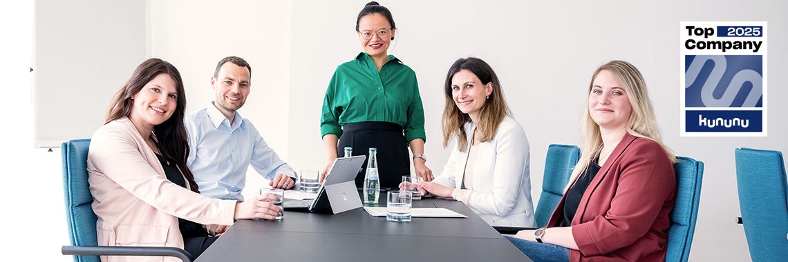 Fünf Mitarbeitende sitzen und stehen um einen Konferenztisch und lächeln in die Kamera. Auf dem Tisch liegen Laptops und Wasserflaschen. Rechts oben ist das „Top Company 2025“-Siegel von Kununu zu sehen.