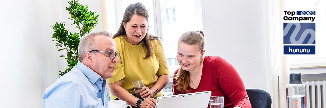 Zwei Frauen und ein Mann schauen gemeinsam lächelnd auf einen Laptop. Sie sitzen an einem Tisch mit Getränken, im Hintergrund steht eine Pflanze. Rechts oben ist das „Top Company 2025“-Siegel von Kununu zu sehen.