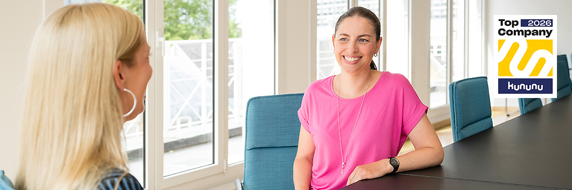 Zwei Frauen sitzen sich an einem Konferenztisch gegenüber und unterhalten sich freundlich. Rechts oben ist das „Top Company 2026“-Siegel von Kununu zu sehen