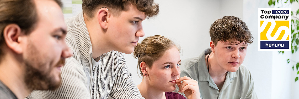 Vier junge Mitarbeitende schauen konzentriert auf einen Bildschirm. Rechts oben ist das „Top Company 2026“-Siegel
