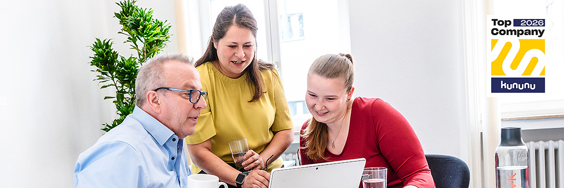 Zwei Frauen und ein Mann schauen gemeinsam lächelnd auf einen Laptop. Sie sitzen an einem Tisch mit Getränken, im Hintergrund steht eine Pflanze. Rechts oben ist das „Top Company 2026“-Siegel von Kununu zu sehen.
