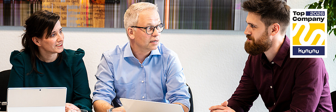 Zwei Männer und eine Frau sitzen an einem Tisch im Gespräch, vor ihnen liegen Tablets. Rechts oben ist das „Top Company 2026“-Siegel von Kununu zu sehen.