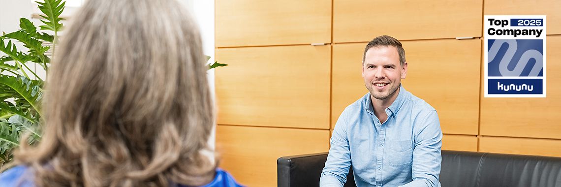 Ein Mann im hellblauen Hemd sitzt auf einem Sofa und spricht mit einer Frau. Im Hintergrund sind Holzverkleidung und Pflanzen zu sehen. Rechts oben befindet sich das „Top Company 2025“-Siegel von Kununu.