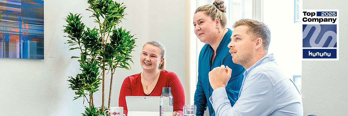 Drei Mitarbeitende unterhalten sich freundlich an einem Tisch mit Laptop und Getränken. Im Hintergrund steht eine große Pflanze. Rechts oben ist das „Top Company 2025“-Siegel von Kununu zu sehen.