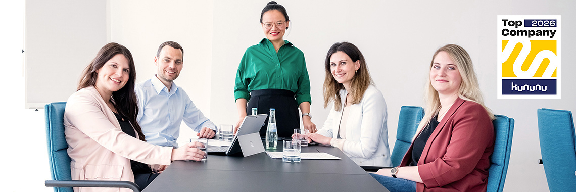 Fünf Mitarbeitende sitzen und stehen um einen Konferenztisch und lächeln in die Kamera. Auf dem Tisch liegen Laptops und Wasserflaschen. Rechts oben ist das „Top Company 2026“-Siegel von Kununu zu sehen.