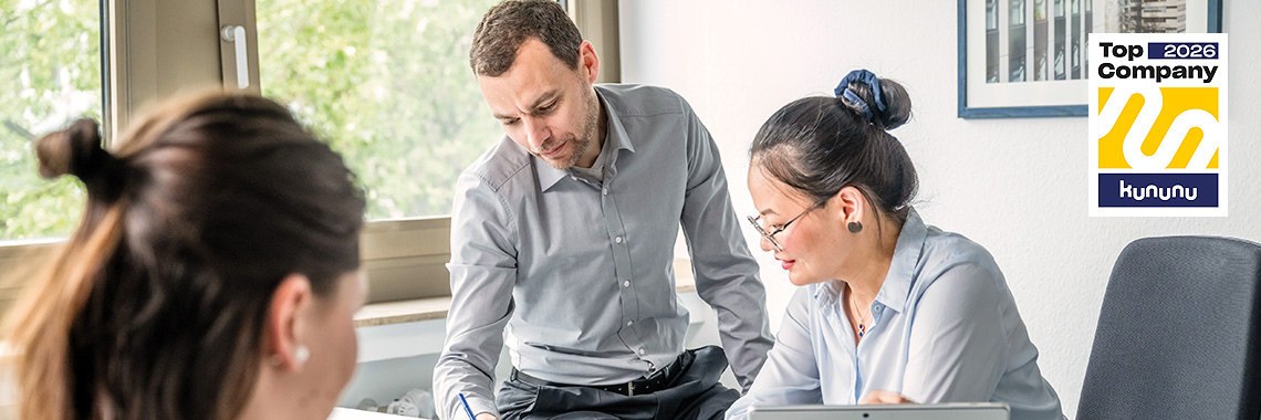 Drei Mitarbeitende arbeiten gemeinsam an einem Tisch, zwei von ihnen schauen auf Unterlagen. Rechts oben ist das „Top Company 2026“-Siegel von Kununu zu sehen.
