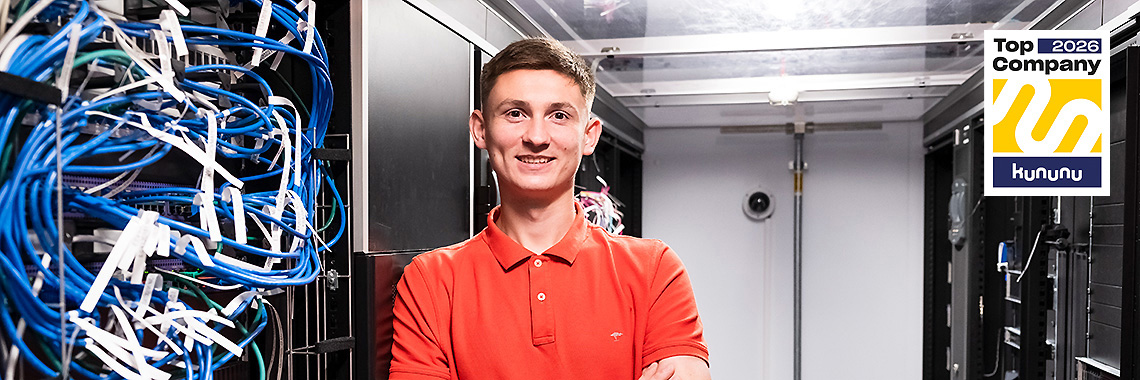 Ein junger Mann im roten Poloshirt steht lächelnd in einem Serverraum mit sichtbaren Netzwerkkabeln. Oben rechts ist das „Top Company 2026“-Siegel von Kununu zu sehen.